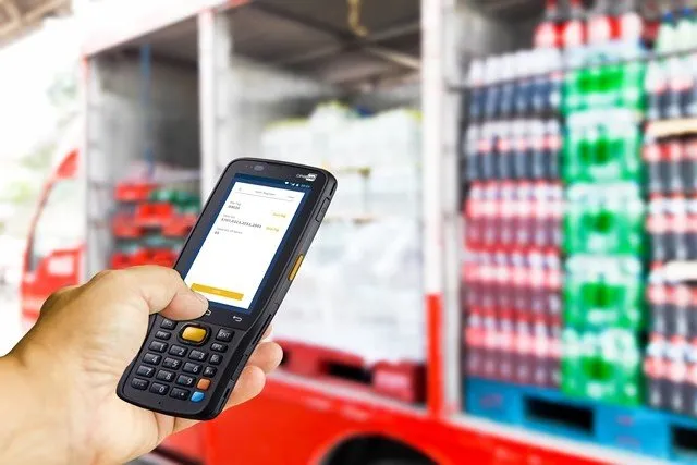 A hand holds a mobile device displaying a screen, with a beverage truck stocked with drinks in the background.